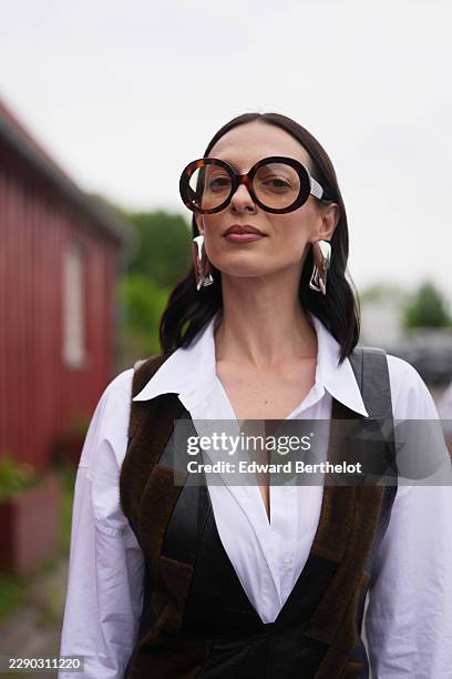 Guest wears long straight dark brown hair with a center part, oversized round black sunglasses with dark lenses, silver large earrings, a white shirt...