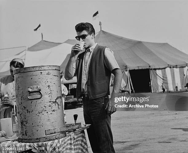 American musician and actor Ricky Nelson, wearing a dark-coloured waistcoat over a light-coloured short-sleeve shirt and sunglasses, drinking from a...