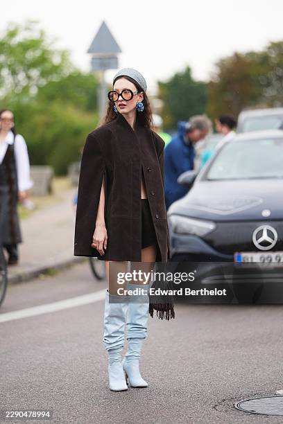 Guest wears a gray knit hat, long wavy brown hair, oversized round tortoiseshell sunglasses with tinted lenses, blue earrings, a dark brown cape...