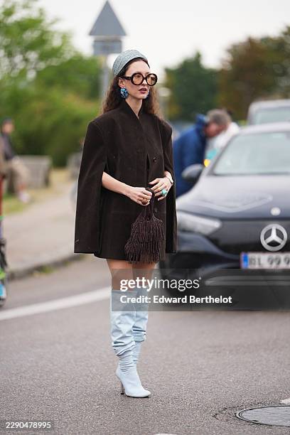 Guest wears a gray knit hat, long wavy brown hair, oversized round tortoiseshell sunglasses with tinted lenses, blue earrings, a dark brown cape...