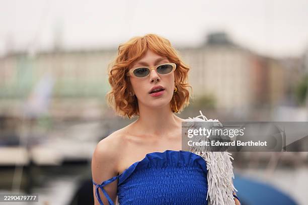 Guest wears short wavy red hair, beige oval sunglasses with dark lenses, lipstick, a white fringe stole, a cobalt blue strapless oversized ruffled...