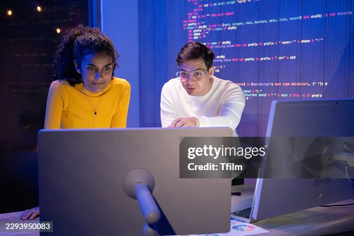 Two Young Programmers Collaborate Late, Reviewing Source Code on Computer Monitor in Dark Office. Colleagues Focus on Screen with Code Projected on Wall Behind Them During Night Session. Two Young Programmers Collaborate Late, Reviewing Source Code on Computer Monitor in Dark Office. Colleagues Focus on Screen with Code Projected on Wall Behind Them During Night Session.