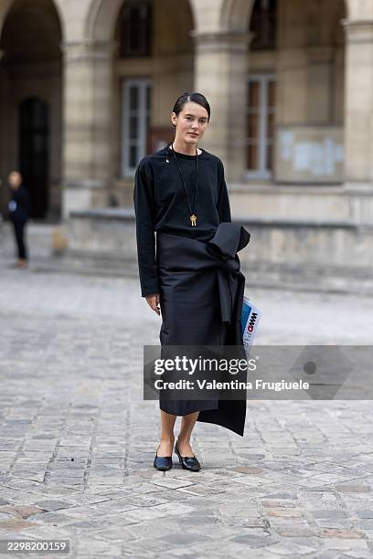 Guest wears a black sweater, a black and gold necklace, a black midi pencil skirt with a bow on the side and black leather pumps outside Carven show...