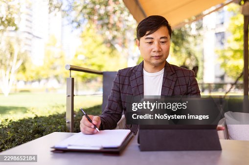 Asian man writing notes while using tablet outdoors Asian man writing notes while using tablet outdoors