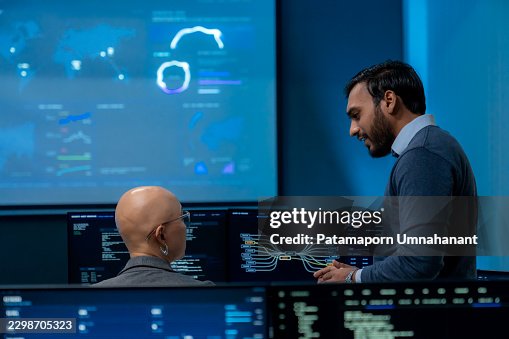Male Software Engineer Explaining Cybersecurity Code Logic to Colleague Working at Computer Workstation in Control Center Male Software Engineer Explaining Cybersecurity Code Logic to Colleague Working at Computer Workstation in Control Center