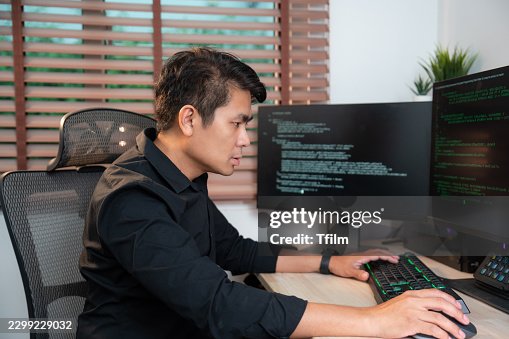 Computer Programmer Typing Code on Keyboard While Focusing on Dual Monitor Setup Displaying Source Code. IT Professional Working at Desk in Modern Home Office, Wearing Black Shirt. Computer Programmer Typing Code on Keyboard While Focusing on Dual Monitor Setup Displaying Source Code. IT Professional Working at Desk in Modern Home Office, Wearing Black Shirt.