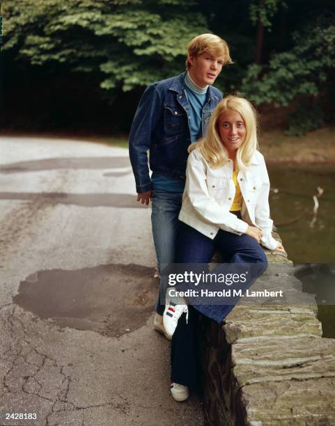 Young man in denim jacket and jeans stands outdoors behind a seated blonde girl in white denim jacket, jeans, and sneakers, circa 1970.