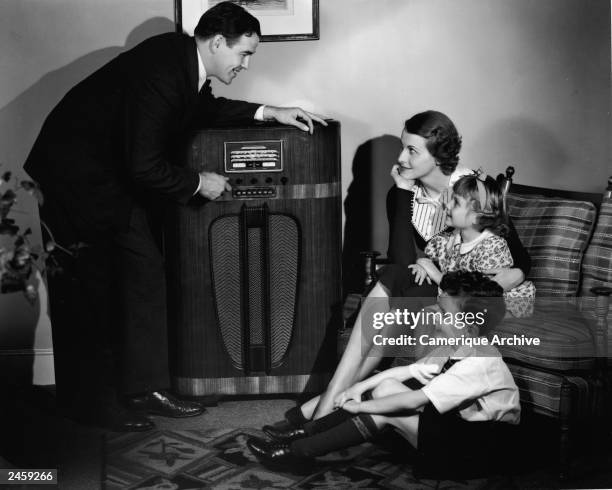 Father adjusts the tuning dial on a radio console as his family sits and listens in their living room, 1930s.