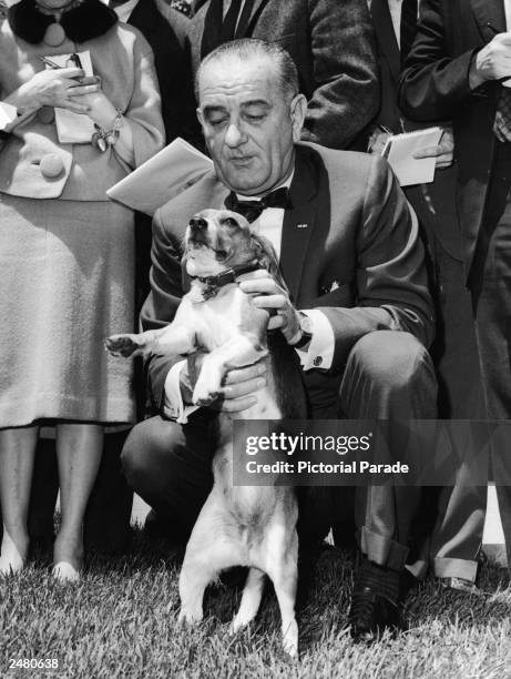 President Lyndon Baines Johnson kneels and pets his pet beagle 'Him,' as reporters look on on the White House lawn, Washington, DC, 1964.