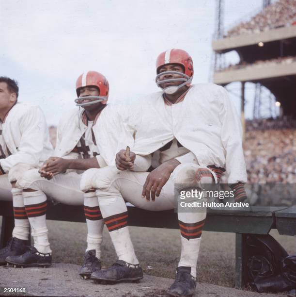 American football player Jim Brown sits on the bench during a game, wearing his helmet and uniform for the Cleveland Browns, circa 1960.