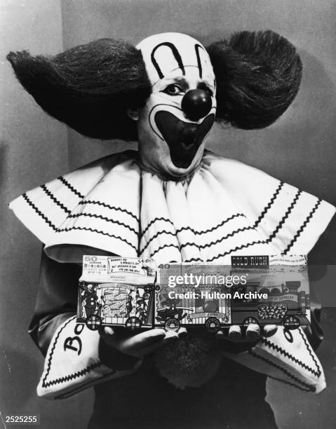 Promotional portrait of Bozo the Clown holding boxes of Bozo Express Bazooka bubble gum with a surprised expression on his face, circa 1965.