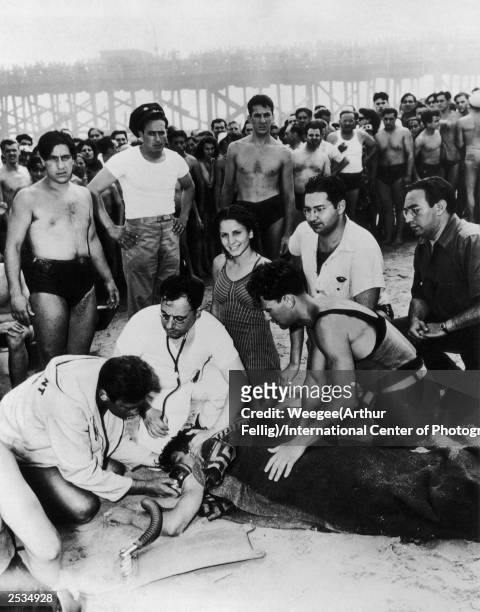 Life guard and a doctor attempt to save a swimmers life on Coney Island Beach, New York, 1940. A woman oblivious to the trauma smiles at the camera...