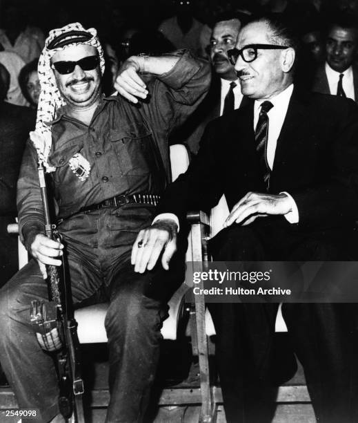 Yasser Arafat , leader of the PLO , holding a Russian-made machine gun, laughs while sitting next to Jordanian premier Abdul Mon'er Rifai during the...