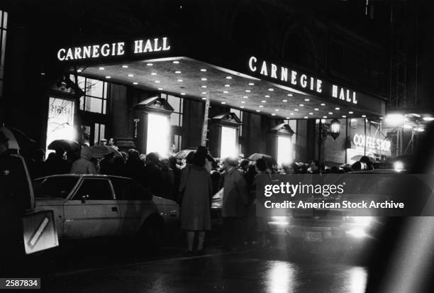 Crowd of people with umbrellas stand in front of Carnegie Hall at night, New York City, circa 1975.