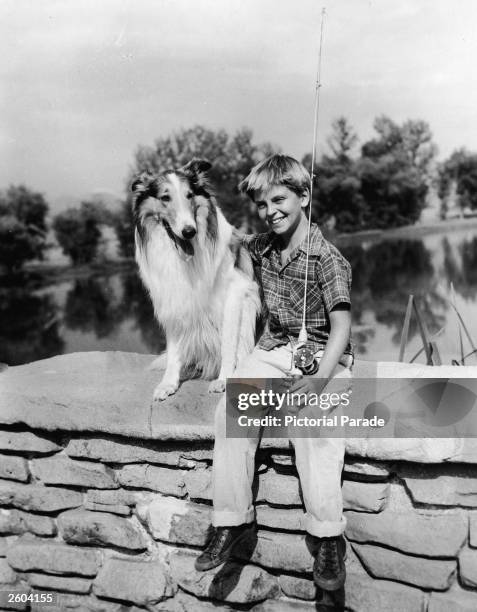 American child actor Tommy Rettig , holding a fishing pole, sits on the stone wall of a bridge with his arm around Lassie in a promotional portrait...