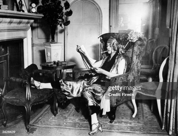 Lady wearing a smoking suit, reading a newspaper by the fireplace.