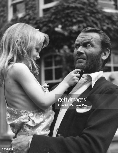 English actor Sir John Mills, wearing a beard for the role of 'Scott, of the Antarctic', holding his young daughter Juliet who is attempting to shave...
