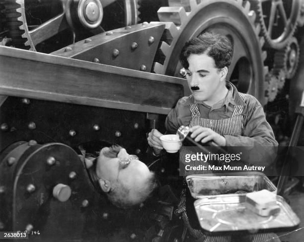 British comedian and director Charlie Chaplin pours a drink from a flask for Chester Conklin , who is imprisoned by a piece of industrial machinery...