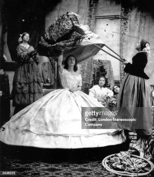 Woman wearing a crinoline being dressed with the aid of long poles to lift her dress over the hoops.
