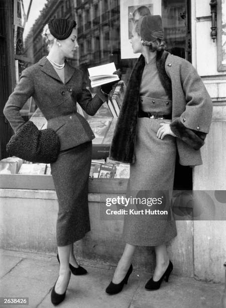 Two models standing outside a bookshop wearing tweed suits from French designer Pierre Balmain's autumn/winter collection - a red suit with black...