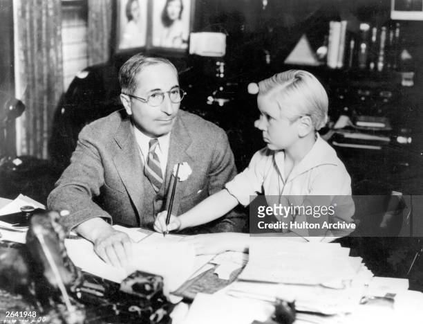 Child actor Jackie Cooper signing a piece of paper while seated with MGM Vice President Louis B Mayer circa 1928.