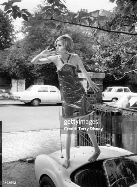 Twiggy modelling a transparent plastic halterneck dress, standing on the boot of a car.