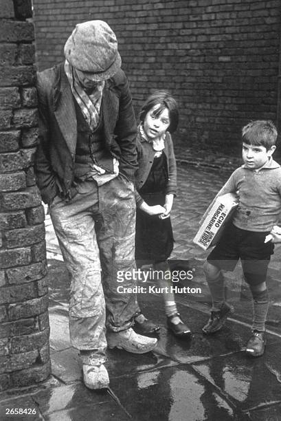 An unemployed man leaning against a wall in Wigan, with two children looking on, November 1939. Original Publication: Picture Post - 228 - Wigan -...