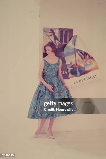 Woman modelling a blue summer dress stands in front of posters advertising holidays abroad.