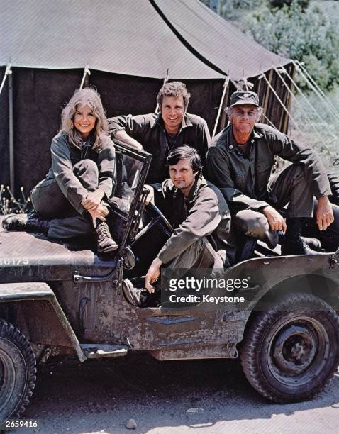 American actor, director and writer Alan Alda in the driving seat of a jeep, surrounded by Loretta Swit and other cast members of the hit television...