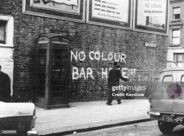 Black man walking down a Notting Hill Street, where race riots had recently taken place. The graffiti behind him reads, 'No Colour Bar Here - Yet'.
