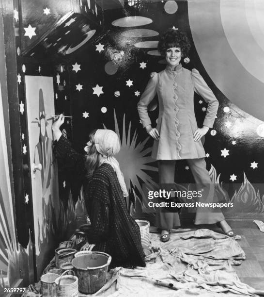 Model Paulene Stone wearing a velvet coat from the Apple boutique, standing in front of a night sky backdrop, while a set designer paints a figure on...