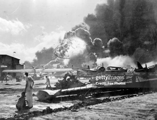 An explosion at the Naval Air Station, Ford Island, Pearl Harbour during the Japanese attack. Sailors stand amid wrecked watching as the USS Shaw...