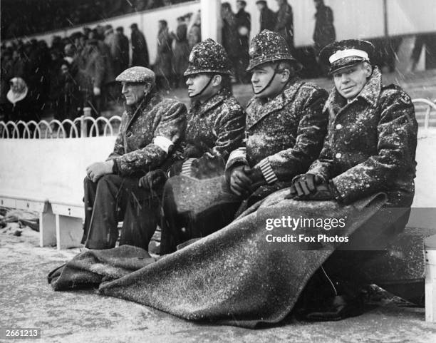 Policemen and ambulancemen sitting on the touchline at White Hart Lane football ground, during a blizzard.