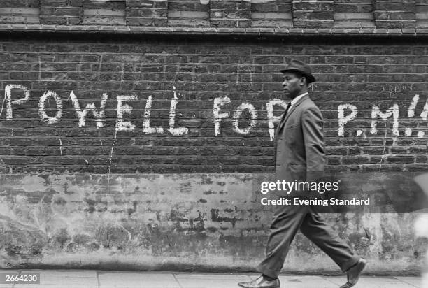 Black man walking past graffiti stating 'Powell For PM' , referring to British Conservative politician Enoch Powell who caused controversy with his...