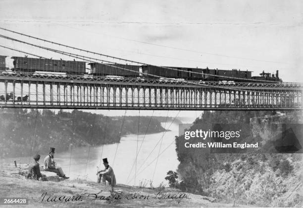 Party of men sit on the Niagara cliffs as a steam train and a horse and carriage cross the river on the two-tier suspension bridge. Built in 1855 by...