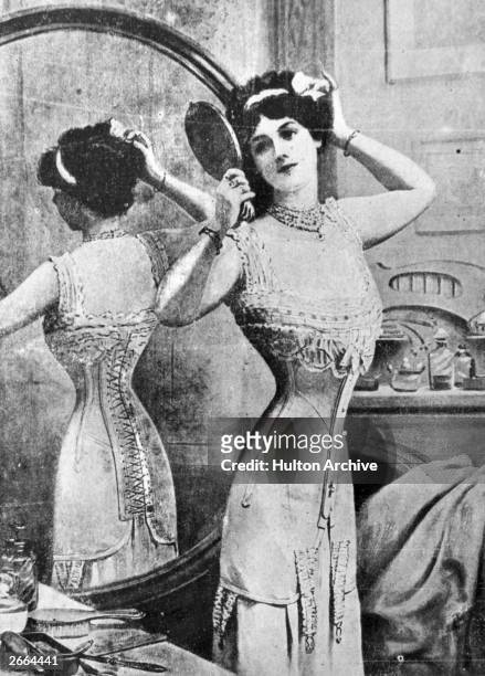 An Edwardian lady, wearing a tightly-laced corset, performs her toilette in front of the mirror. Original Publication: A magazine advertisement for...
