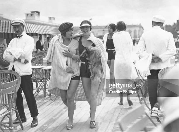 Two women wearing swim suits and shrouded in a blanket, one of them carrying a dog, walking along an open air pier in Deauville, Normandy.