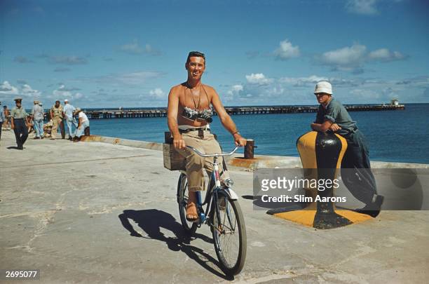 Photographer Slim Aarons riding a Huffy bicycle along the quayside with cameras slung round his neck during the filming of 'Mister Roberts' in...