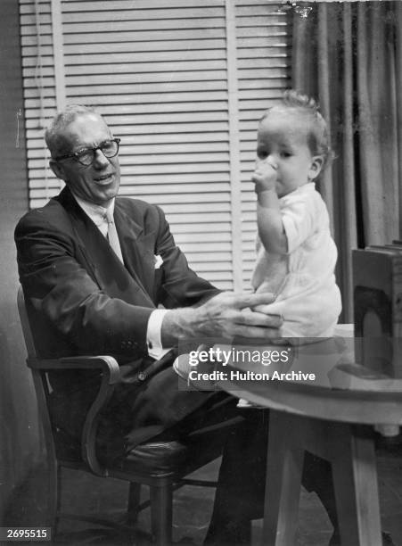 American pediatrician Dr Benjamin Spock , author of, 'The Common Sense Book of Baby and Child Care,' holding onto a baby sitting on a table, circa...