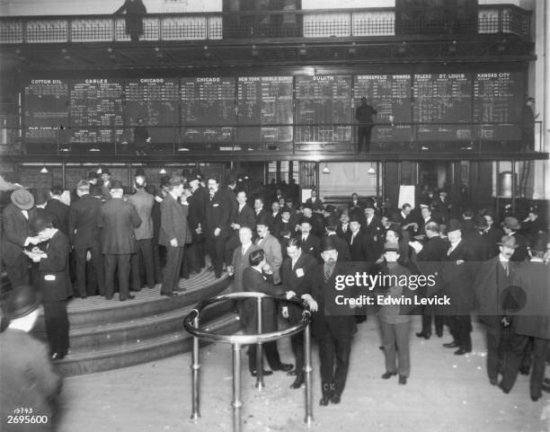 Full-length view of traders on the floor of a stock exchange while a man posts updated figures for commodities such as wheat, cotton oil, corn, oats,...