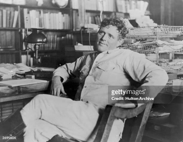 American author Jack London reclines next to his desk in a wooden chair, smiling, his legs crossed.