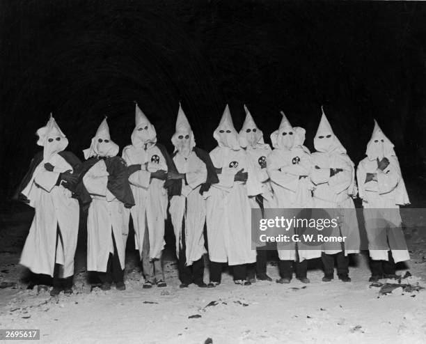 Full-length portrait of Ku Klux Klan members standing in a row with their arms folded in white hoods and robes.