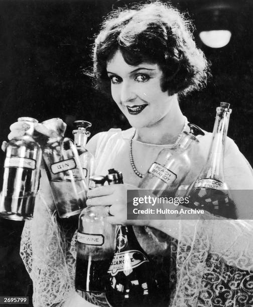 Woman smiles while holding bottles of various types of alcohol, including peach brandy, port wine, gin, absinthe, and forbidden fruit.