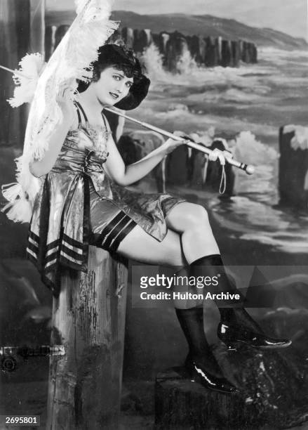 Portrait of a young woman modeling a silk bathing costume, a parasol, patent leather shoes, and black stockings while sitting on a wooden post in...