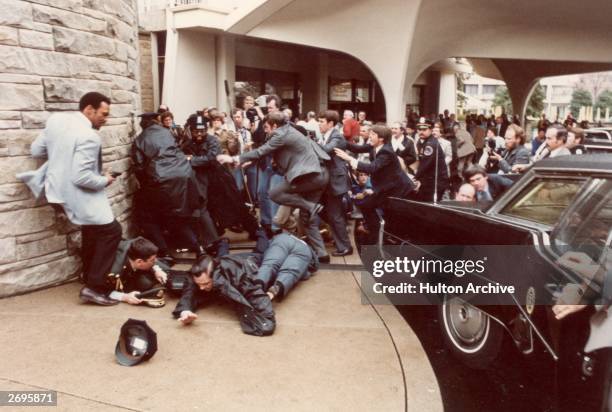 View of police officers and Secret Service agents as they dive to protect President Ronald Reagan amid a panicked crowd during an assassination...