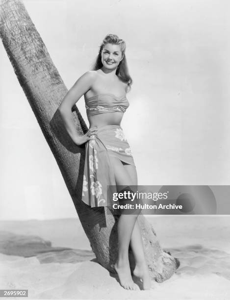 Full-length studio portrait of American actor and swimmer Esther Williams, wearing a bandeau top and sarong with a tropical print, leaning against a...