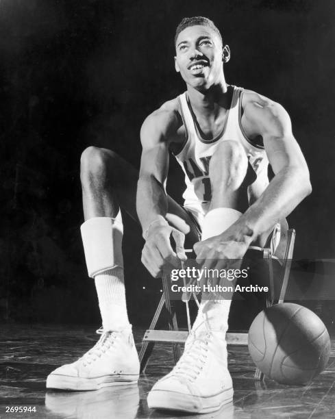 American basketball player Wilt Chamberlain, wearing a University of Kansas uniform, sits in a chair and ties his shoelace. A basketball rests on the...