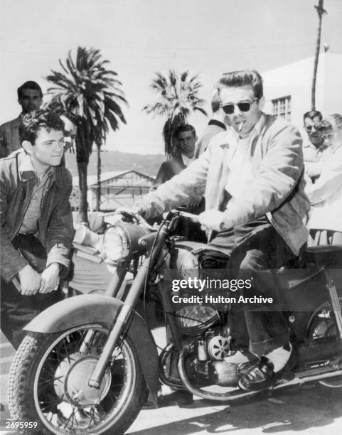 American actor James Dean sits on a motorcycle with a cigarette in his mouth, as young male and female fans look on.