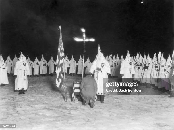 Man kneels at a burning cross as a hooded Klan member stands over him with a chalice during a Ku Klux Klan nighttime initiation ceremony, 1920s....