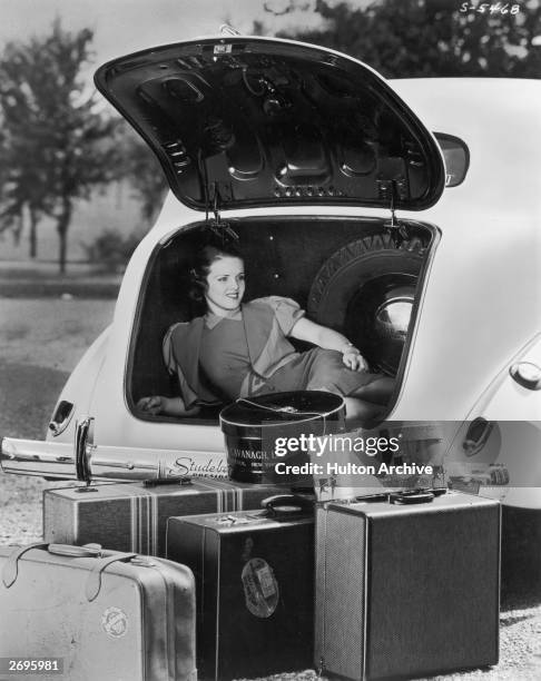 Woman reclines in the trunk of a Studebaker while luggage sits outside the automobile.